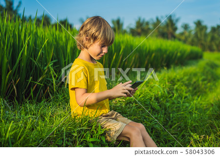 Happy child sitting on the field holding tablet. Boy sitting on the grass on sunny day. Home 58043306