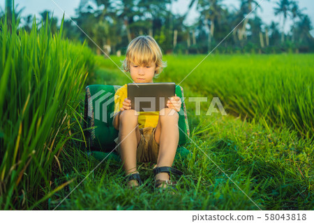Happy child sitting on the field holding tablet. Boy sitting on the grass on sunny day. Home 58043818