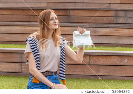 Young beautiful female worker holding paper lunch bag sitting on a wooden bench in a park. Lunch 58064885