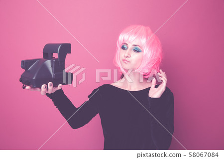 Young stylish woman holding retro camera and standing in a pink wig in a bright studio. 58067084