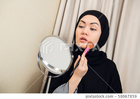 Arab woman applying makeup on her face, wearing traditional Arabian dress Arab woman applying makeup on her face, wearing traditional Arabian dress 58067859