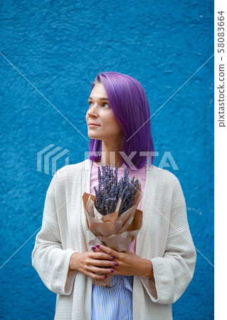 Purple haired woman in white cardigan holding a lavender bouquet in her hands. 58083664