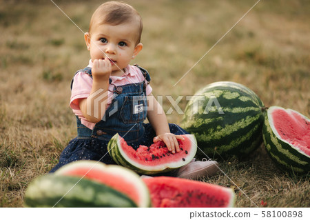 Cute little baby girl laughing with watermelons. Girl dressed in denim dress. Summer portrait of 58100898