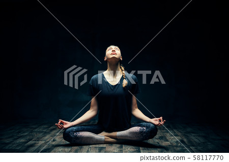 Young attractive woman practicing yoga sitting in lotus pose and meditating in dark room 58117770