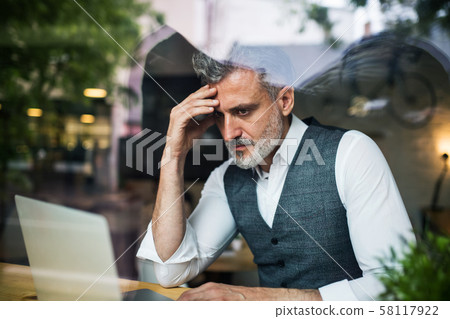 Serious mature man sitting at the table in a cafe, using laptop. Serious mature man sitting at the table in a cafe, using laptop. 58117922