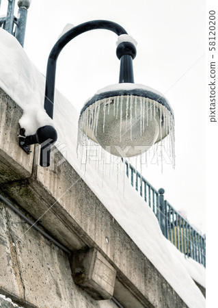 Outdoor street lantern hanged on concrete wall covered with icicles on cold winter day. Winter 58120200