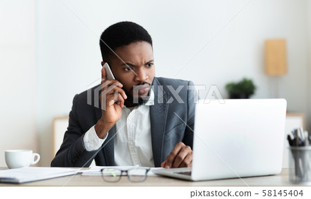 Concerned businessman talking on cellphone and looking at laptop screen Concerned businessman talking on cellphone and looking at laptop screen 58145404