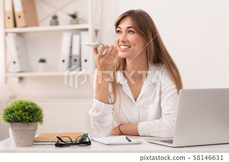 Woman Using Smartphone Voice Assistant Sitting At Laptop In Office Woman Using Smartphone Voice Assistant Sitting At Laptop In Office 58146631