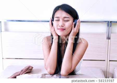 Cheerful young woman listen music with headphones while sitting on the bed with pillow, she is enjoying on relax time. Lifestyle relaxation concept. Cheerful young woman listen music with headphones while sitting on the bed with pillow, she is enjoying on relax time. Lifestyle relaxation concept. 58153597