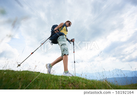 Woman hiker hiking on grassy hill, wearing backpack, using trekking sticks in the mountains Woman hiker hiking on grassy hill, wearing backpack, using trekking sticks in the mountains 58192164