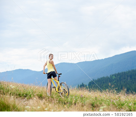 Attractive female cyclist with yellow mountain bicycle, enjoying sunny day in the mountains Attractive female cyclist with yellow mountain bicycle, enjoying sunny day in the mountains 58192165