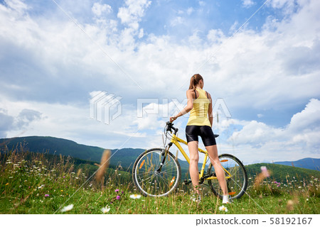 Attractive female cyclist with yellow mountain bicycle, enjoying sunny day in the mountains Attractive female cyclist with yellow mountain bicycle, enjoying sunny day in the mountains 58192167