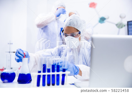 Woman chemist who works at the table and has two colleagues behind her Woman chemist who works at the table and has two colleagues behind her 58221458
