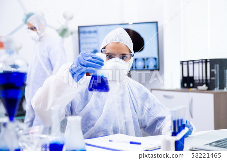 Scientist in safety glasses looks at a beaker with blue liquid and smoke Scientist in safety glasses looks at a beaker with blue liquid and smoke 58221465