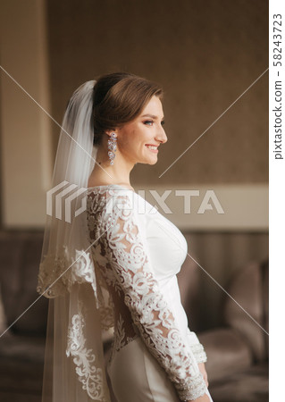 Portrait of beautiful bride standing by the window at home and tying on earrings. Charming bride in 58243723