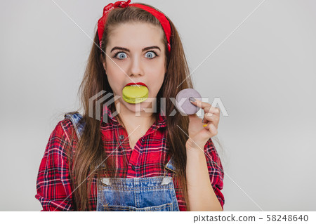 Cheerful young childish girl is holding sweet macaroon in teeth, looking at the camera her eyes Cheerful young childish girl is holding sweet macaroon in teeth, looking at the camera her eyes 58248640