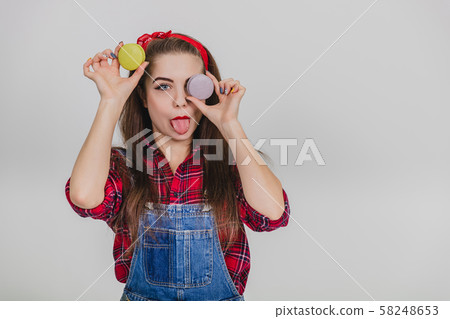 Beautiful happy cute young pretty woman holding two macaroons, hiding her eye behind blue one Beautiful happy cute young pretty woman holding two macaroons, hiding her eye behind blue one 58248653