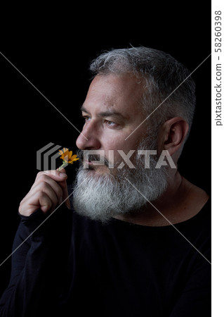 Portrait of a brutal gray haired man with a beard sniffing yellow flowers on a black background 58260398