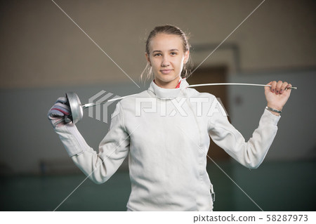 A young smiling woman fencer standing in the gym with a sword behind her shoulders A young smiling woman fencer standing in the gym with a sword behind her shoulders 58287973