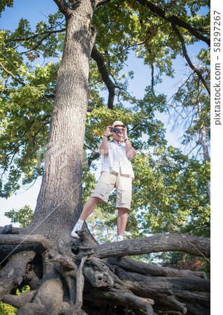 Man wearing shorts standing on big tree and taking photo of forest Man wearing shorts standing on big tree and taking photo of forest 58297267