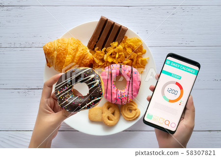 Calories counting and food control concept. woman using Calorie counter application on her smartphone with doughnut in hand and snack , cookies on plate at background 58307821