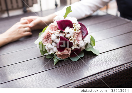 Wedding bouquet on a wooden table. Hands of the newlyweds on the table next to the bouquet. Wedding bouquet on a wooden table. Hands of the newlyweds on the table next to the bouquet. 58315954