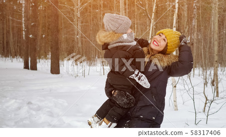 Young mom with little son in her hands is looking at snowfall in winter forest. Young mom with little son in her hands is looking at snowfall in winter forest. 58317406