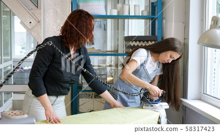 Seamstress ironing clothes with steam, teaching woman iron in tailor workshop. Seamstress ironing clothes with steam, teaching woman iron in tailor workshop. 58317423