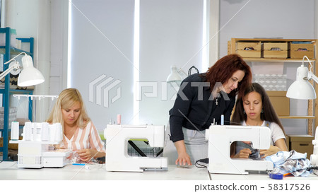 Seamstress teacher teaches her students to sew on sewing machines in workshop. Seamstress teacher teaches her students to sew on sewing machines in workshop. 58317526