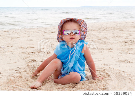 A little girl in blue pareo, sunglasses and a hat sits on the golden sand on the beach by the sea A little girl in blue pareo, sunglasses and a hat sits on the golden sand on the beach by the sea 58359054
