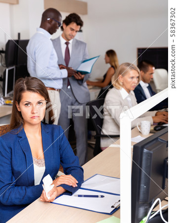 Portrait of frustrated young business woman in coworking space with busy colleagues behind Portrait of frustrated young business woman in coworking space with busy colleagues behind 58367302