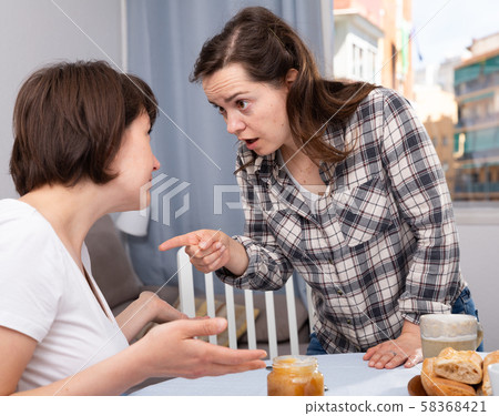Upset adult woman quarrel with female friend at table with cookies Upset adult woman quarrel with female friend at table with cookies 58368421
