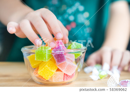 Closeup hands of a little child taking a piece of jelly cube with sugar in a glass bowl. Snacks time, Sugary treats, Party, Kids favorite, Unhealthy, Cavity, Sugar addiction, School, Home, Sweetness. 58369062
