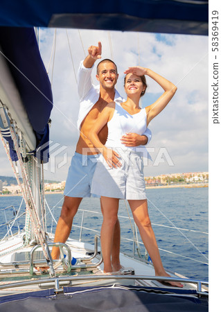 Loving couple standing on deck of sailboat during sea trip along Spain shoreline, looking and pointing to horizon 58369419