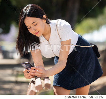 Happy brazilian girl sitting on a bench and using phone in park Happy brazilian girl sitting on a bench and using phone in park 58370497