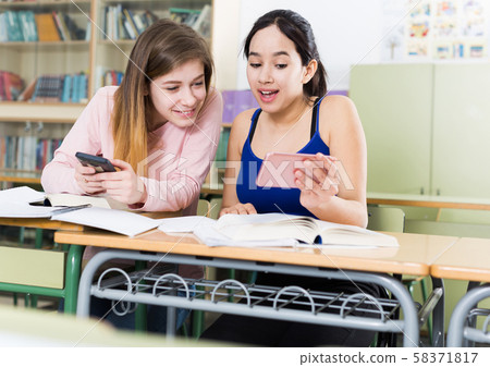 Two girl are sitting at the desk and playing with phones Two girl are sitting at the desk and playing with phones 58371817