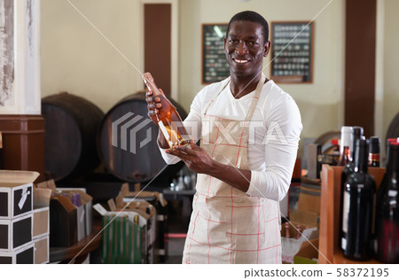 Smiling salesman in apron proposing wine in bottles in winery shop Smiling salesman in apron proposing wine in bottles in winery shop 58372195