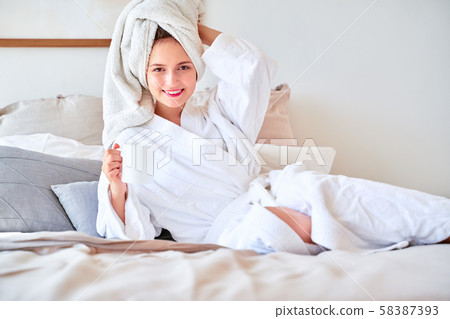 Image of young woman in white bathrobe with mug of tea in her hands lying on bed . Image of young woman in white bathrobe with mug of tea in her hands lying on bed . 58387393