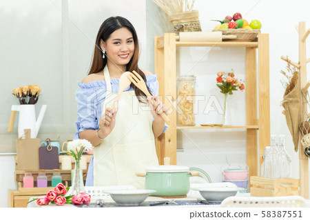 beautiful Asian woman holding wooden cutlery behind dining table with pot , dish and kitchenware in the kitchen room with smiley face 58387551