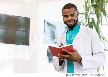 Portrait of a young African man doctor in medical costume, looking at camera, holding red otebook 58390702