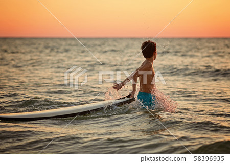 Happy little surfer boy makes splashes with hands at golden sunset 58396935