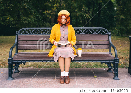 A beautiful red-haired girl with a curly hairstyle is sitting on a park bench 58403603