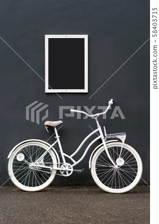 A white painted bicycle stands against a black wall 58403715