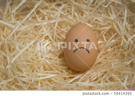 A brown chicken egg with a sad smile lies on a straw in a nest close up 58414395