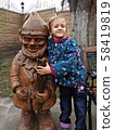 Little happy girl in a green knitted sweater stands next to a wooden gnome 58419819