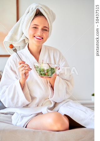 Image of young woman in bathrobe with salad in bowl in her hands sitting on bed. 58437515