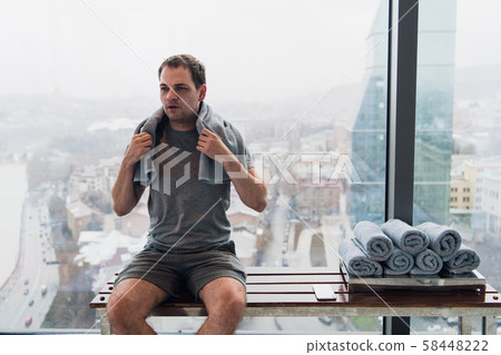 Young, tired man sitting on bench in a gym. Holding mobile phone and towel on his head. Gym at 58448222