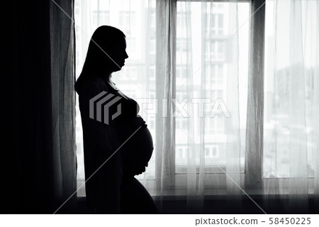 Silhouette of a pregnant woman holding her belly looking out a window with curtains Silhouette of a pregnant woman holding her belly looking out a window with curtains 58450225