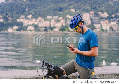 The theme of tourism and travel in Italy. A male cyclist uses a phone on the shore of Lake Como 58466582