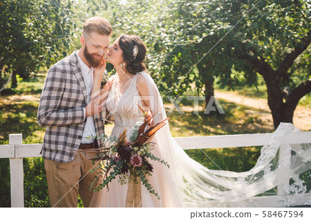 Caucasian couple in love bride and groom standing in embrace near wooden white, rural fence in park 58467594
